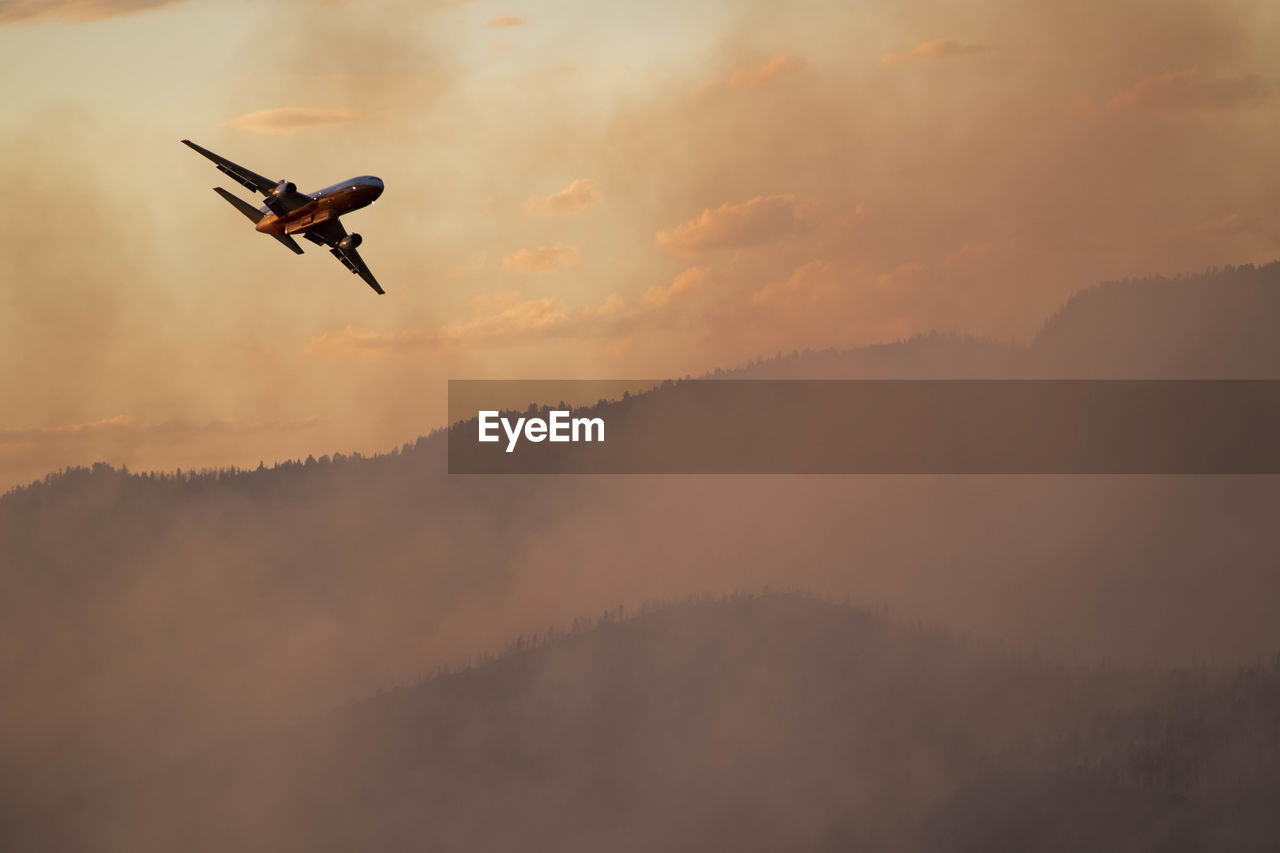 Military aircraft flying over smoke emitting from wildfire in forest during sunset