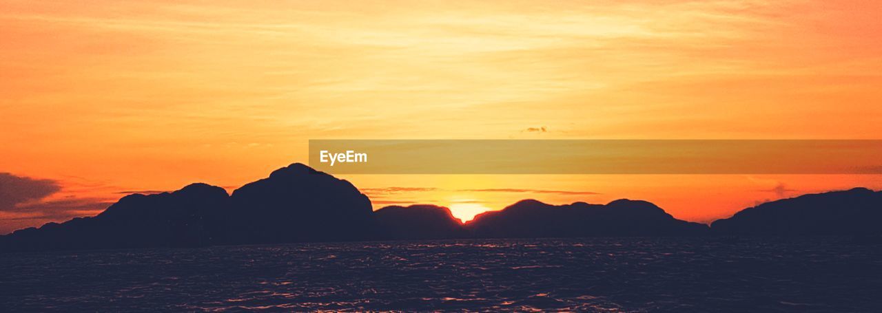 SCENIC VIEW OF SEA BY SILHOUETTE MOUNTAINS AGAINST ORANGE SKY