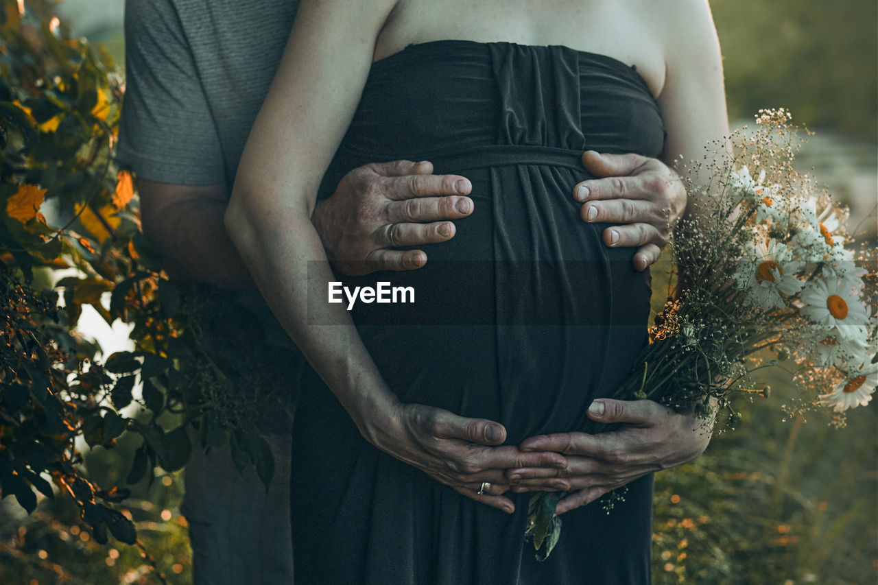 Pregnant woman and her husband hugging her tummy standing outdoors surrounded by nature. pregnancy,