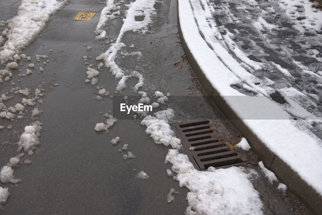 Surface drain on the corner of a tarmac road, surrounded by melting snowfall. winter environment