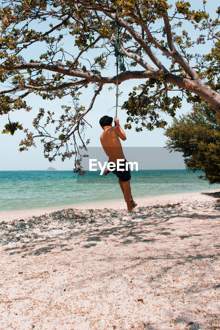 REAR VIEW OF SHIRTLESS BOY ON BEACH AGAINST SKY