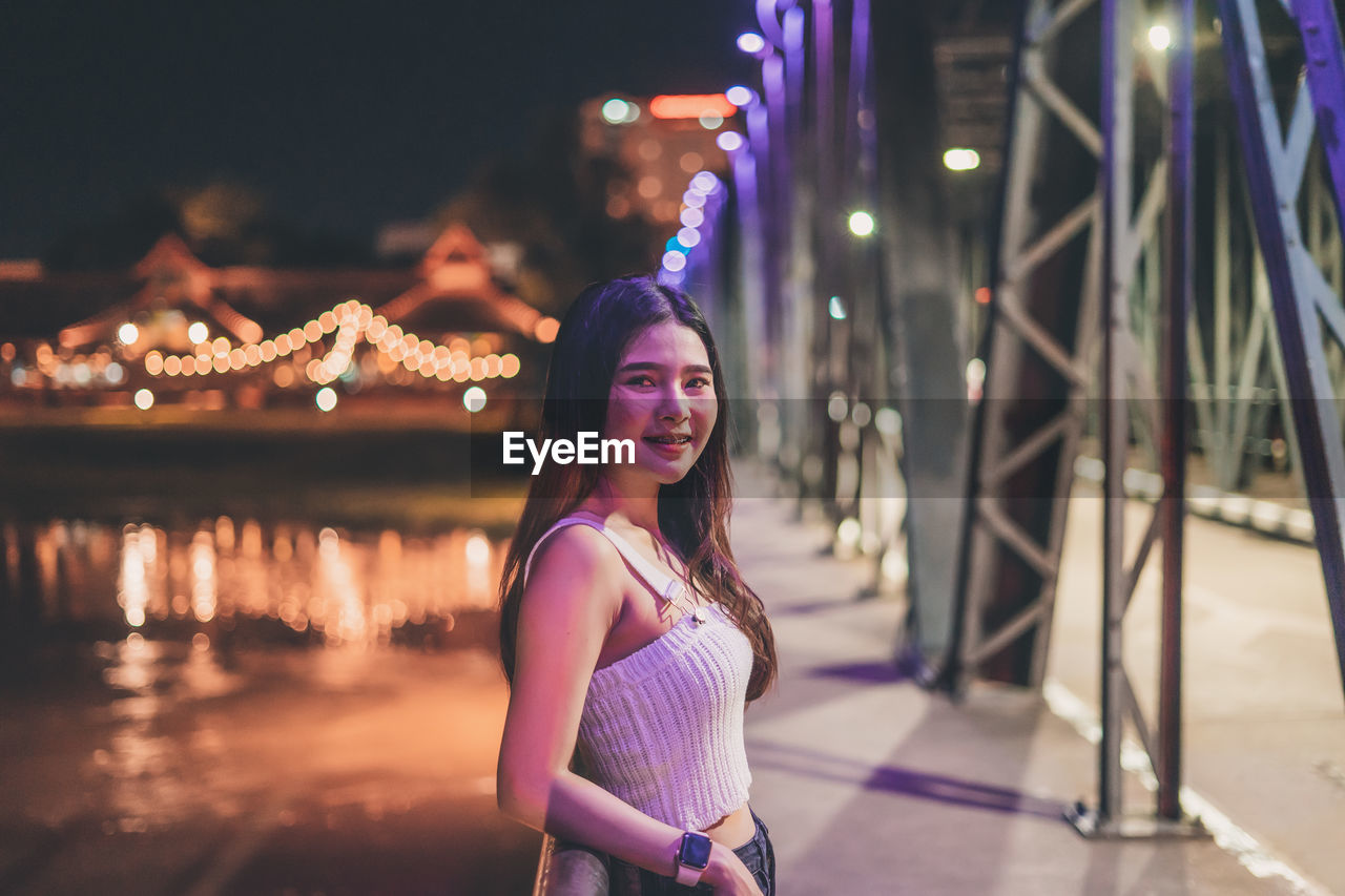 Portrait of smiling young woman standing on bridge over river at night