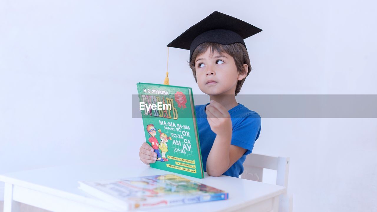 Thoughtful boy in mortarboard holding books at home