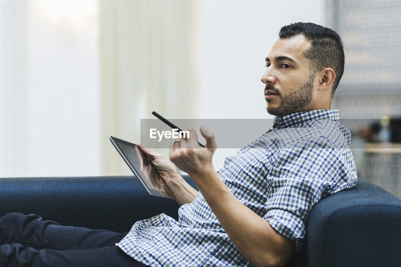 Side view of businessman holding digital tablet while relaxing on sofa at office