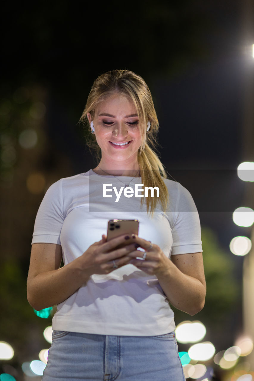 wireless technology, one person, technology, mobile phone, adult, smartphone, portable information device, women, holding, smiling, standing, communication, young adult, happiness, casual clothing, front view, telephone, blond hair, emotion, three quarter length, portrait, night, lifestyles, person, hairstyle, using phone, outdoors, activity, illuminated, long hair, city, portability, waist up, cheerful, leisure activity, focus on foreground, clothing, female, looking, looking at camera