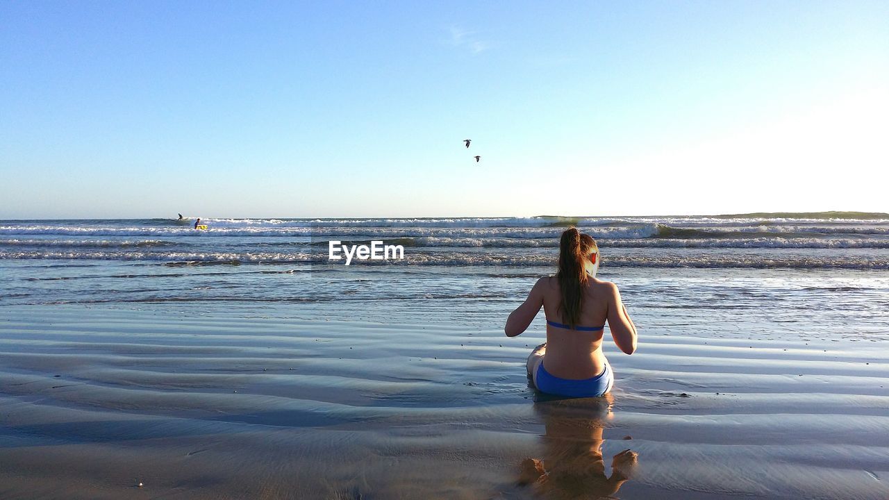 Rear view of woman in bikini sitting on shore at beach against sky