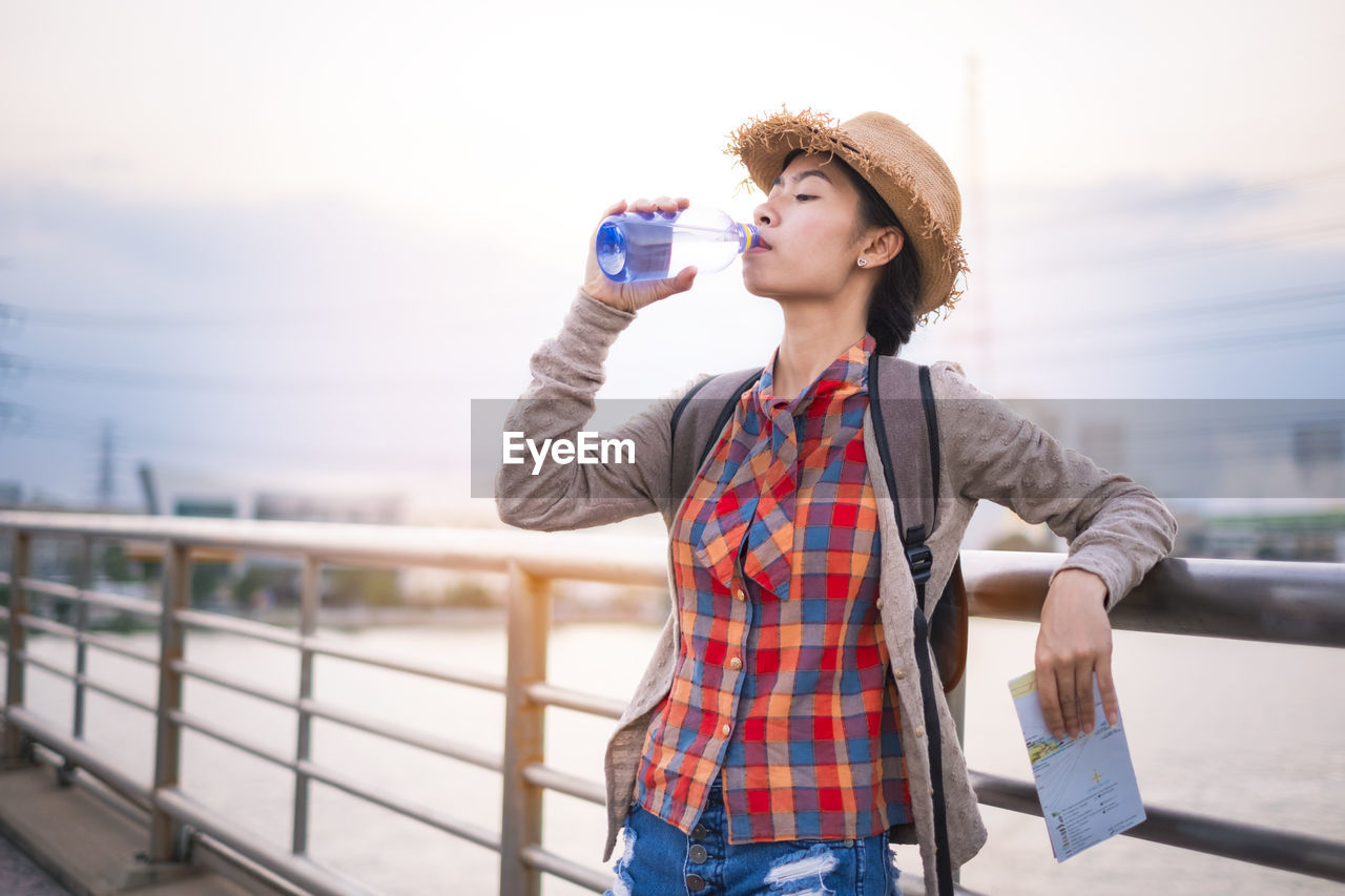 Young woman drinking water while standing against railing and sky