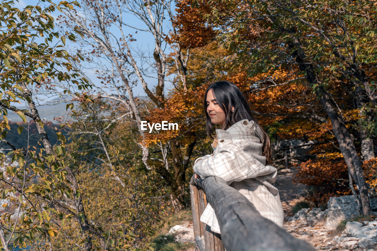 autumn, tree, one person, plant, nature, leaf, leisure activity, women, young adult, woodland, forest, adult, lifestyles, land, winter, wilderness, spring, sitting, day, long hair, clothing, casual clothing, sunlight, beauty in nature, hairstyle, outdoors, tranquility, smiling, relaxation, looking, warm clothing, female, plant part, portrait, three quarter length, full length, growth, person, non-urban scene, happiness