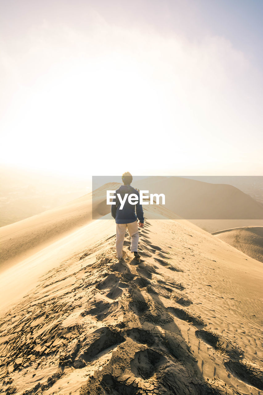 Rear view of man standing on sand dune against sky during sunset