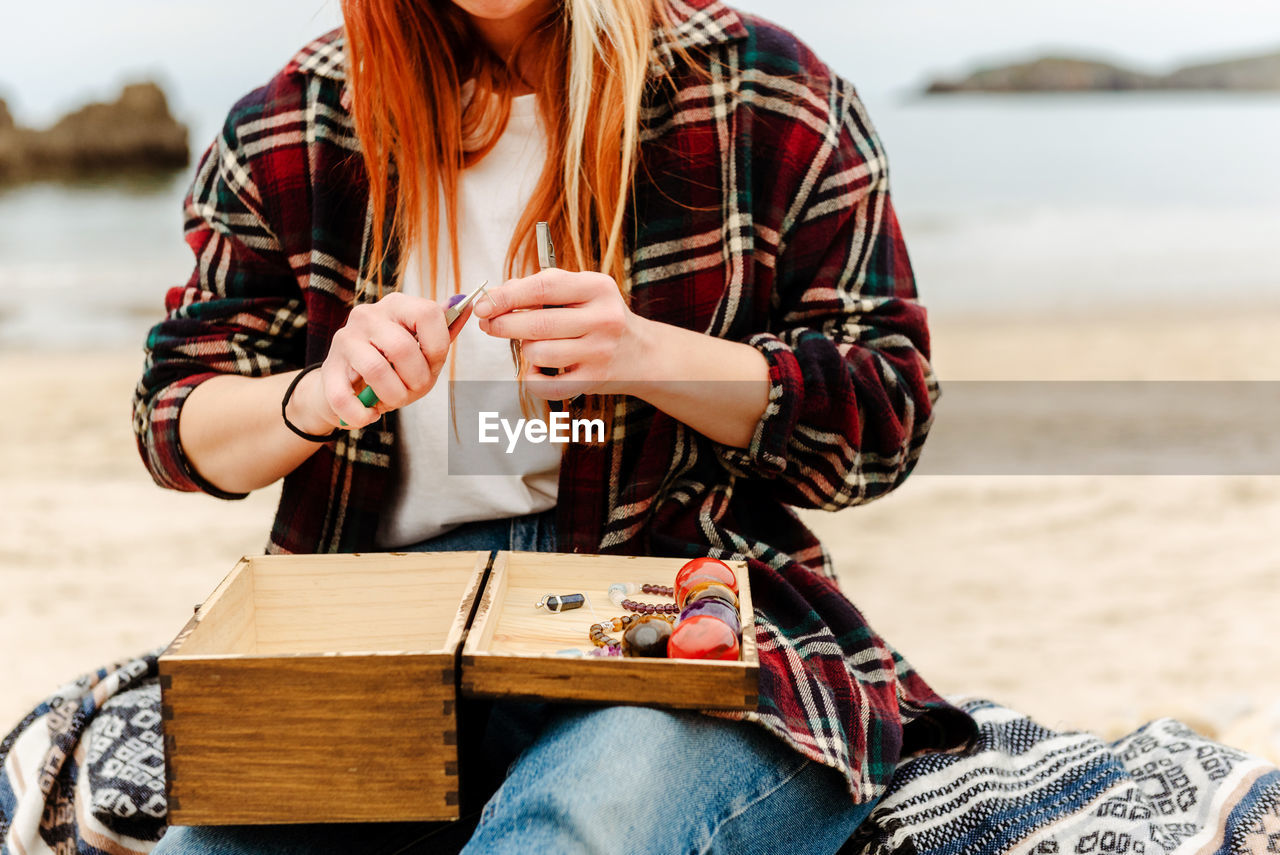 Crop anonymous craftswoman creating handmade bijouterie while using semiprecious stones and sitting on beach near sea