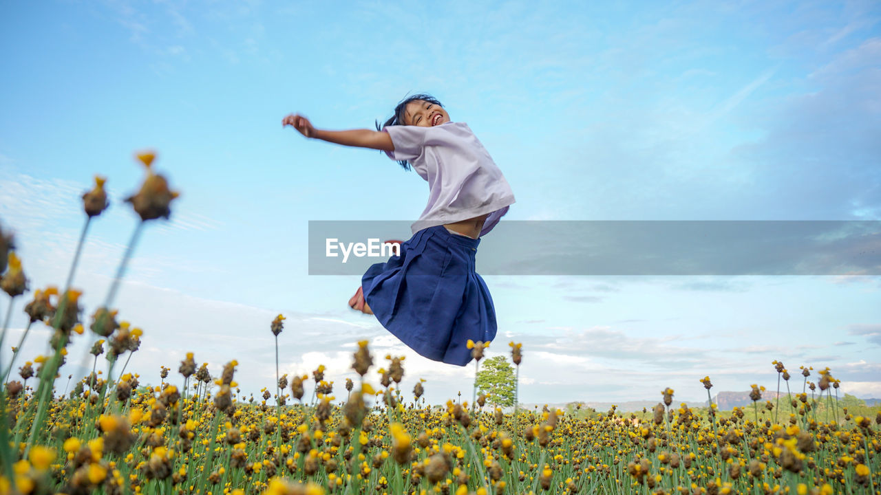 Full length of girl jumping over flowering plants against sky
