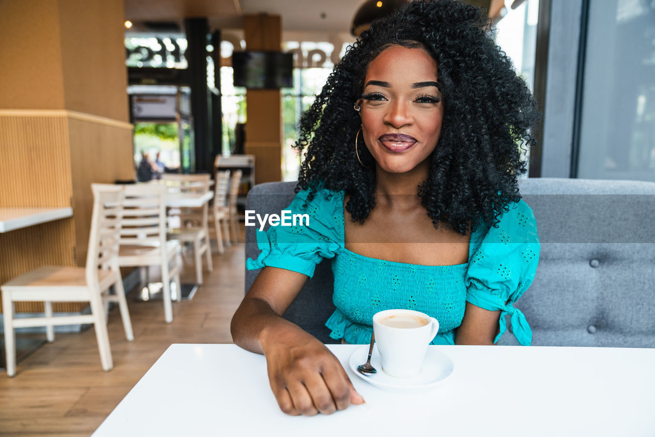 Content african american lady with makeup sitting at table with cup of coffee and looking at camera in cafe