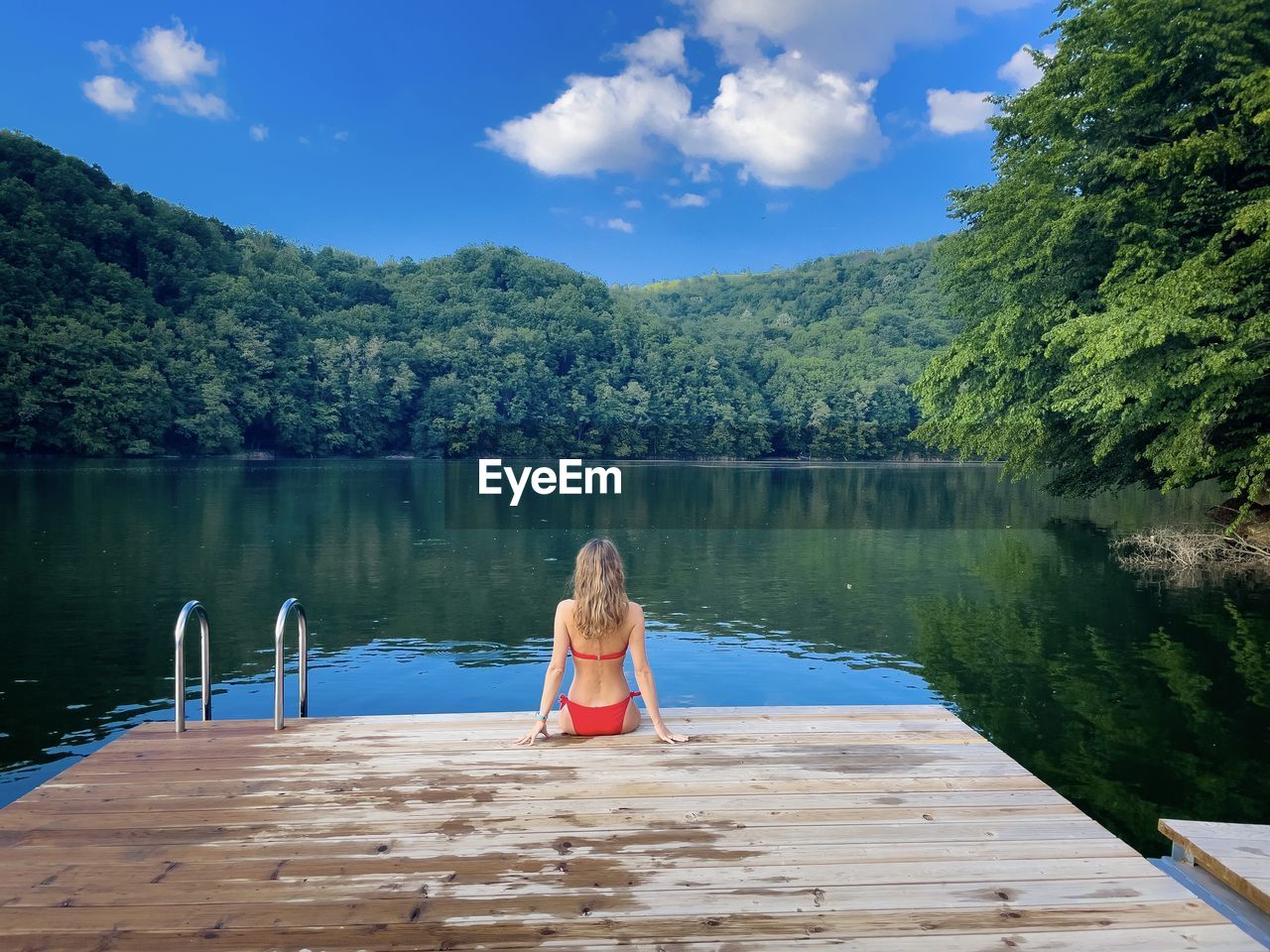 Woman in red bikini sitting down on a wooden pontoon looking at the lake surrounded by green forest