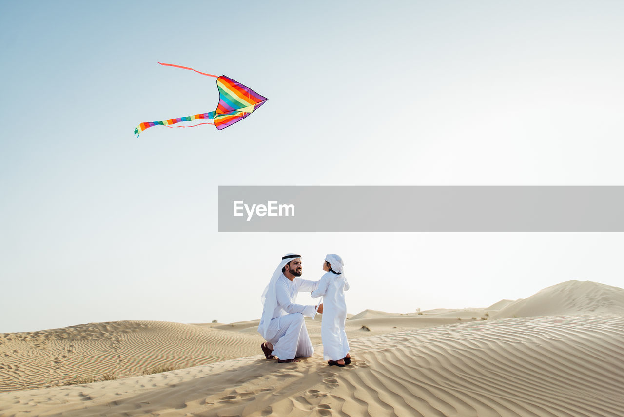 MAN AND WOMAN STANDING ON SAND DUNE AGAINST SKY