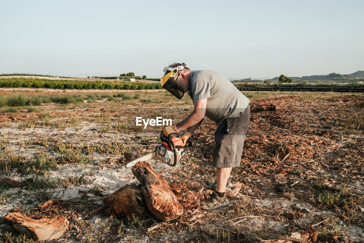 Side view of mature man standing in field and starting modern chainsaw while getting ready for cutting tree trunk in rural area in highlands