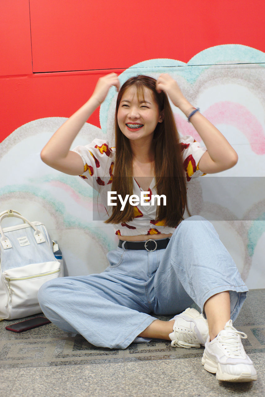 PORTRAIT OF SMILING GIRL SITTING ON BED AGAINST RED WALL