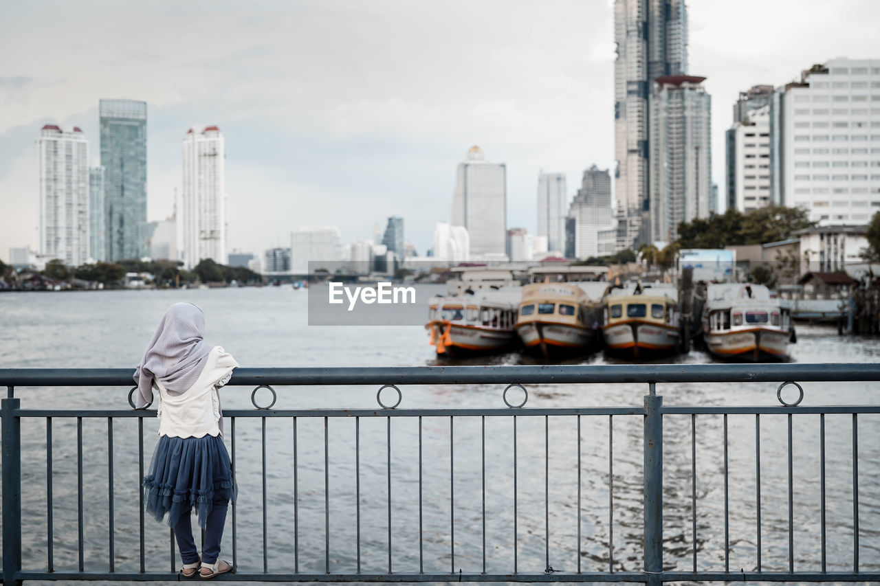 Rear view of a girl standing on railing against buildings of waterfront city