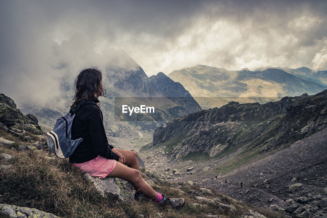 Side view of woman sitting on mountain against cloudy sky