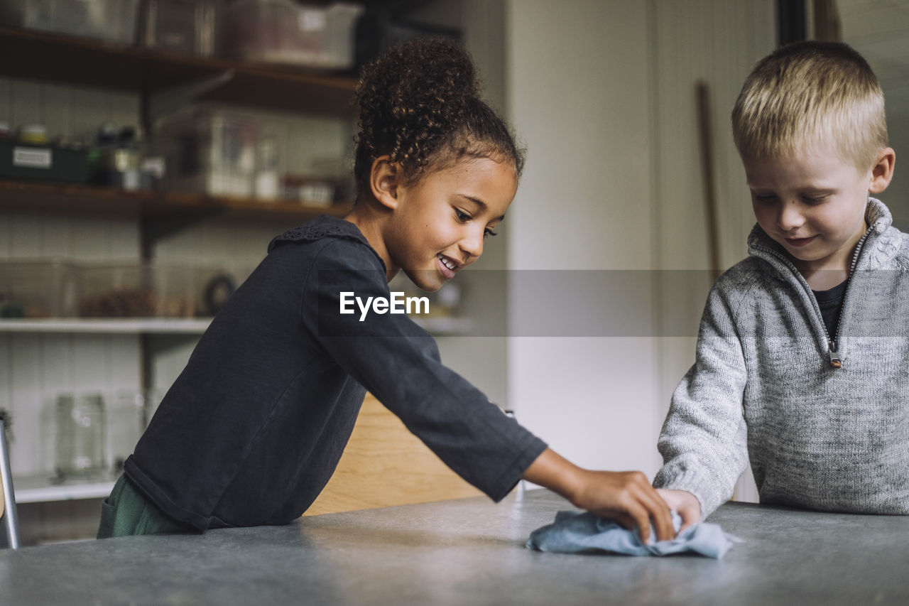 Smiling girl and boy cleaning dining table with napkin at child care center