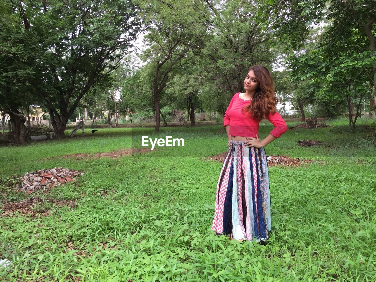 plant, one person, tree, women, grass, green, full length, dress, fashion, nature, young adult, standing, lawn, clothing, flower, hairstyle, front view, lifestyles, adult, field, leisure activity, long hair, day, portrait, land, spring, growth, smiling, looking at camera, casual clothing, outdoors, female, park, beauty in nature, emotion, park - man made space, happiness, looking, red