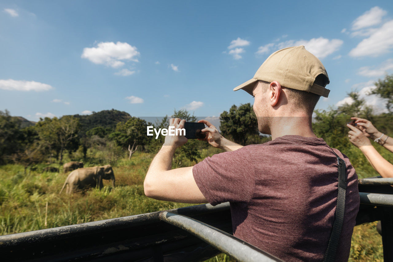 side view of man photographing while sitting on railing against sky