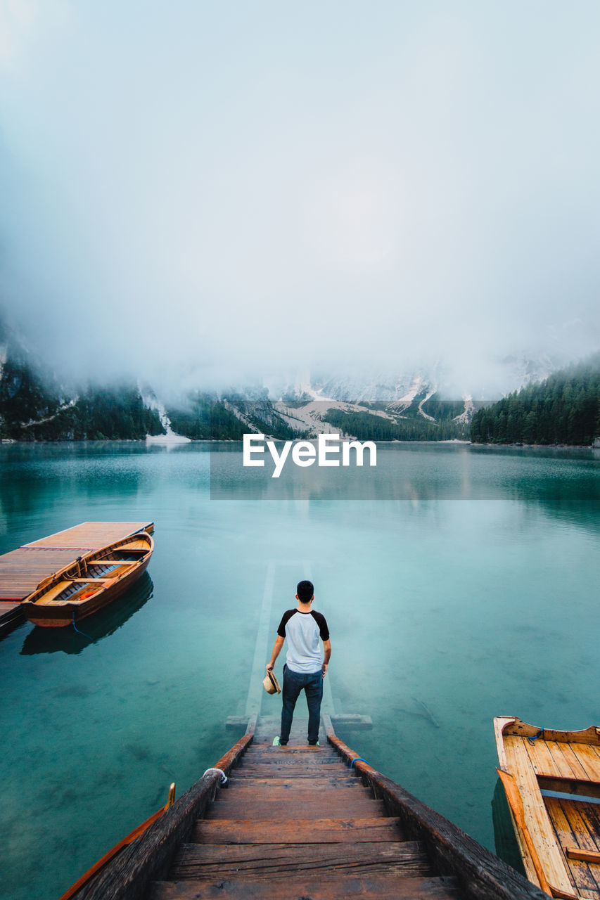 Back view of unrecognizable male traveler standing on wooden quay and admiring amazing scenery of lake with turquoise water in mountains on foggy day