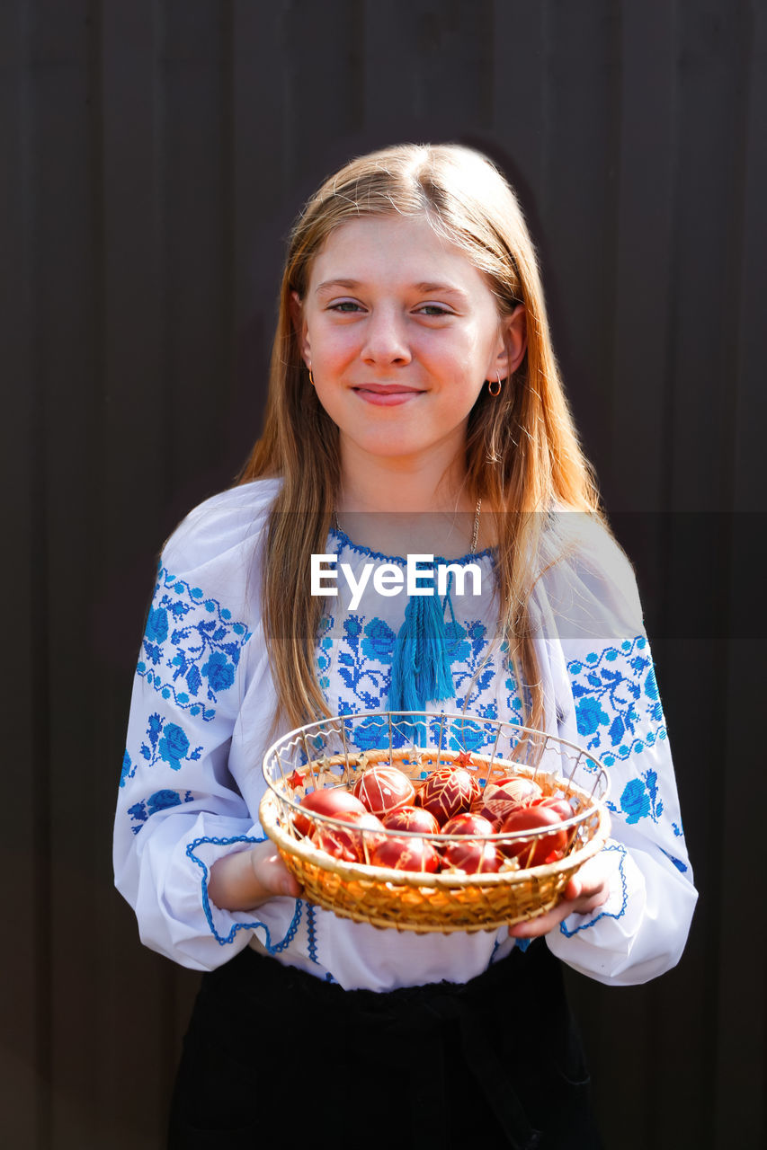 Ukrainian smiling girl in vyshyvanka holding basket of colored red eggs on black background. easter