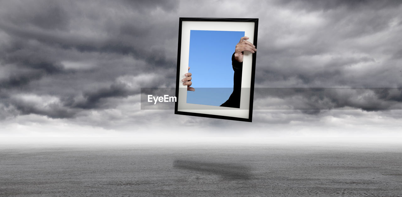 Digital composite image of human hands and picture frame over landscape against storm clouds