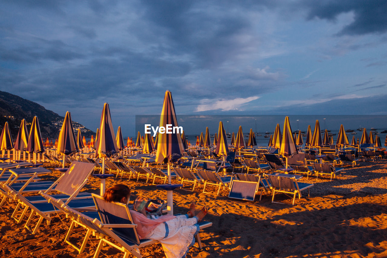 Woman sitting on lounge chair at beach against cloudy sky
