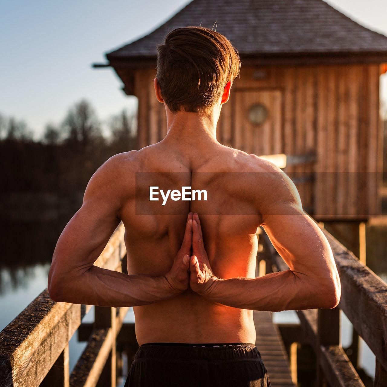 Rear view of shirtless man with hands clasped exercising while standing on pier