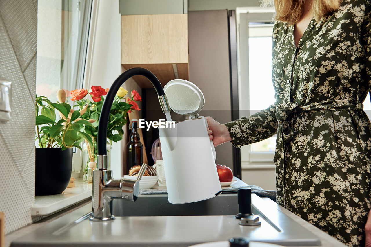 Woman pouring water from faucet into electric kettle at the kitchen
