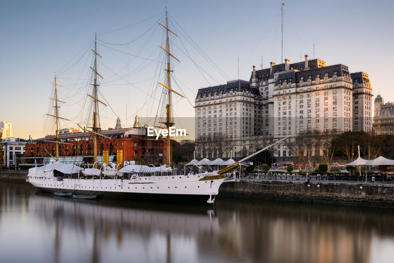 Sailing ship moored in river by buildings against clear sky