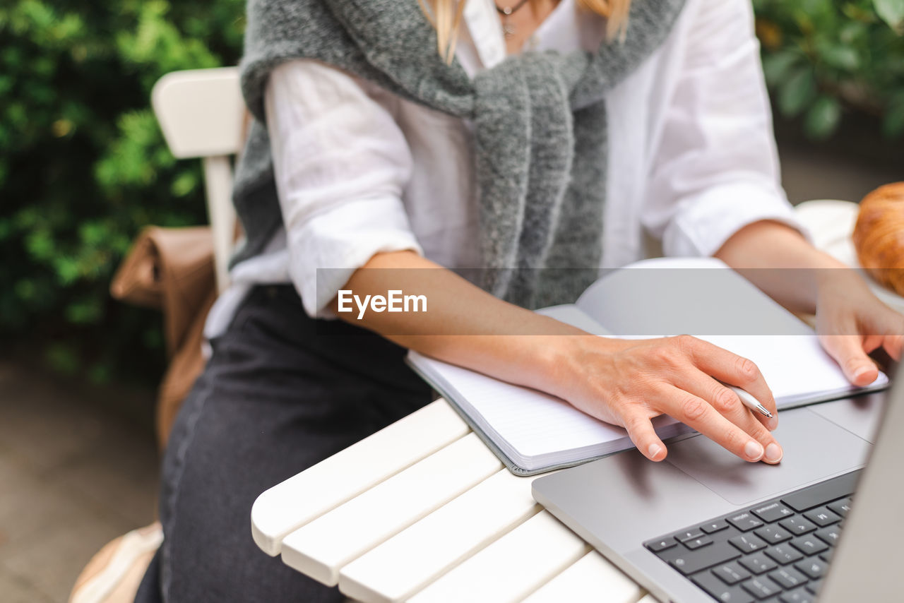 A woman takes notes in front of a laptop during an online business meeting outside.