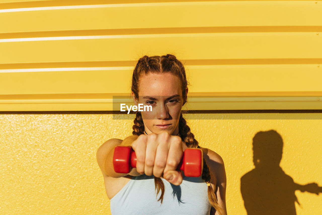 Sportswoman holding dumbbell in front of yellow wall