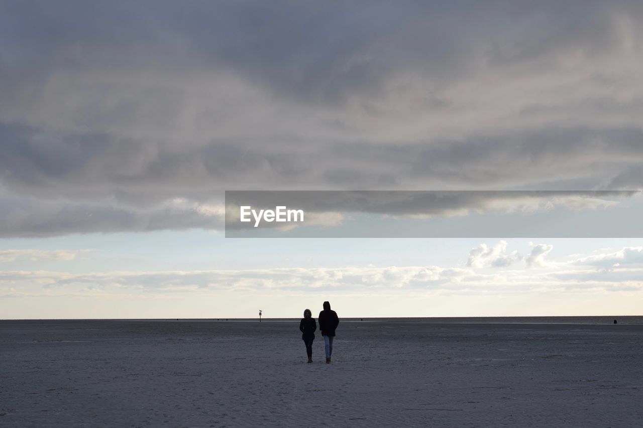 Rear view of friends walking at beach against cloudy sky
