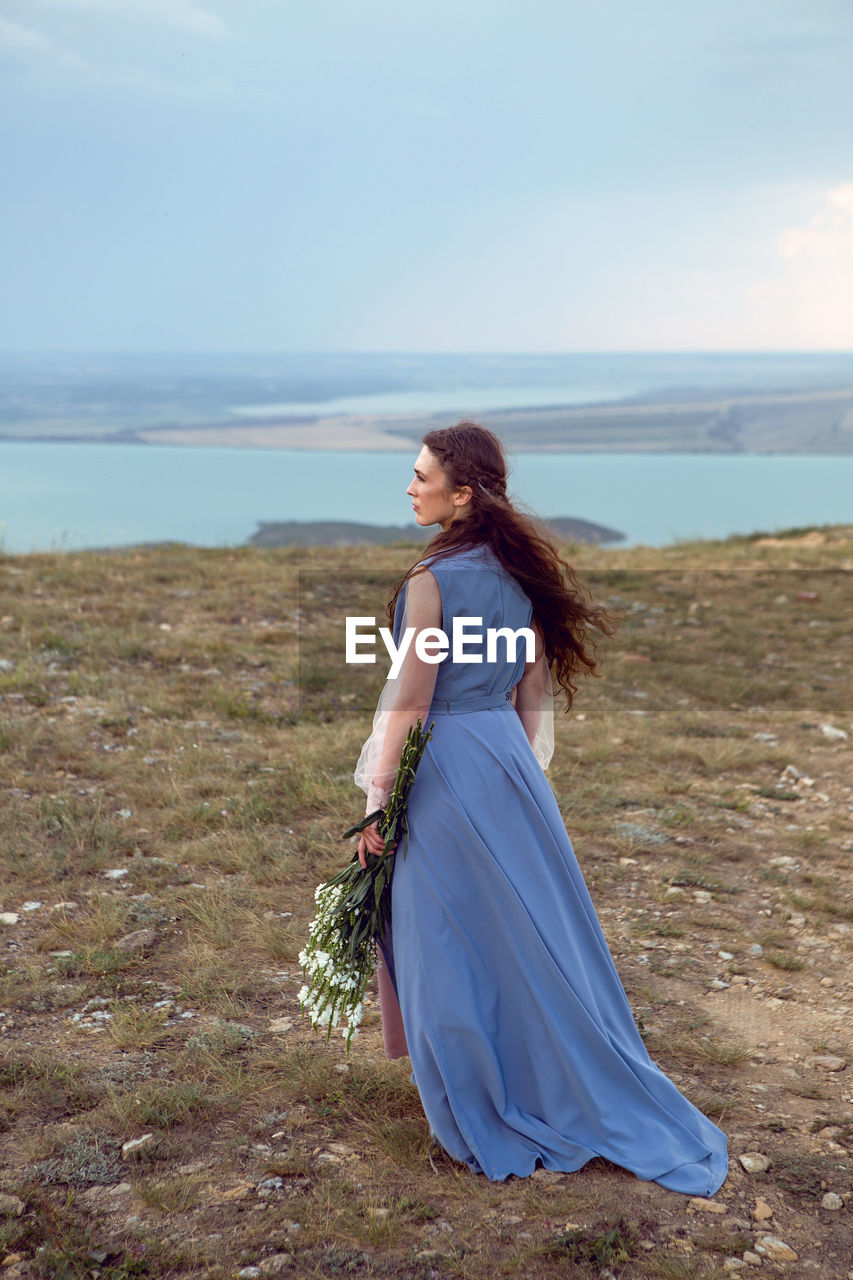 Woman stands on a mountain cliff in a blue long dress in summer