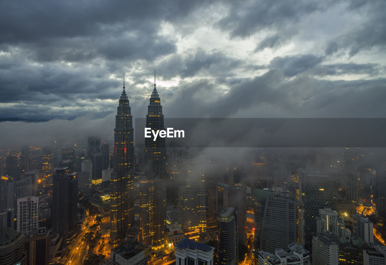 Illuminated buildings in city against cloudy sky