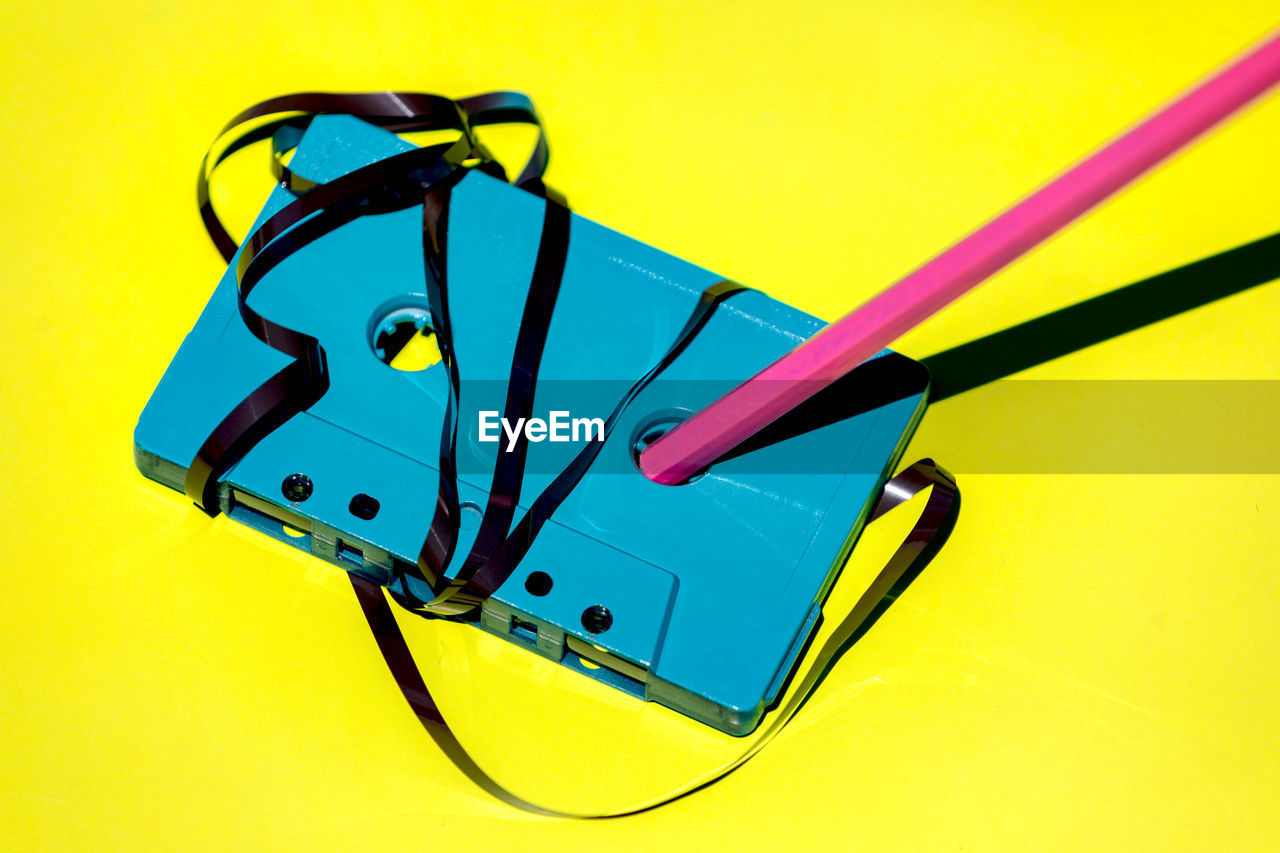 High angle of blue retro cassette with messy magnetic tape and pencil for rewind placed on yellow background in studio