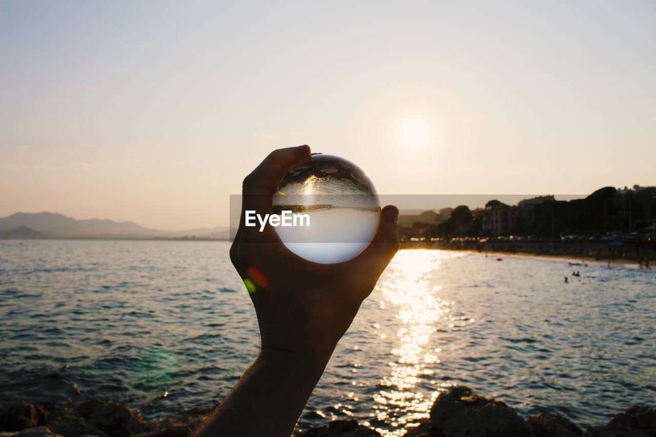 Cropped hand holding crystal ball against sea and sky during sunset