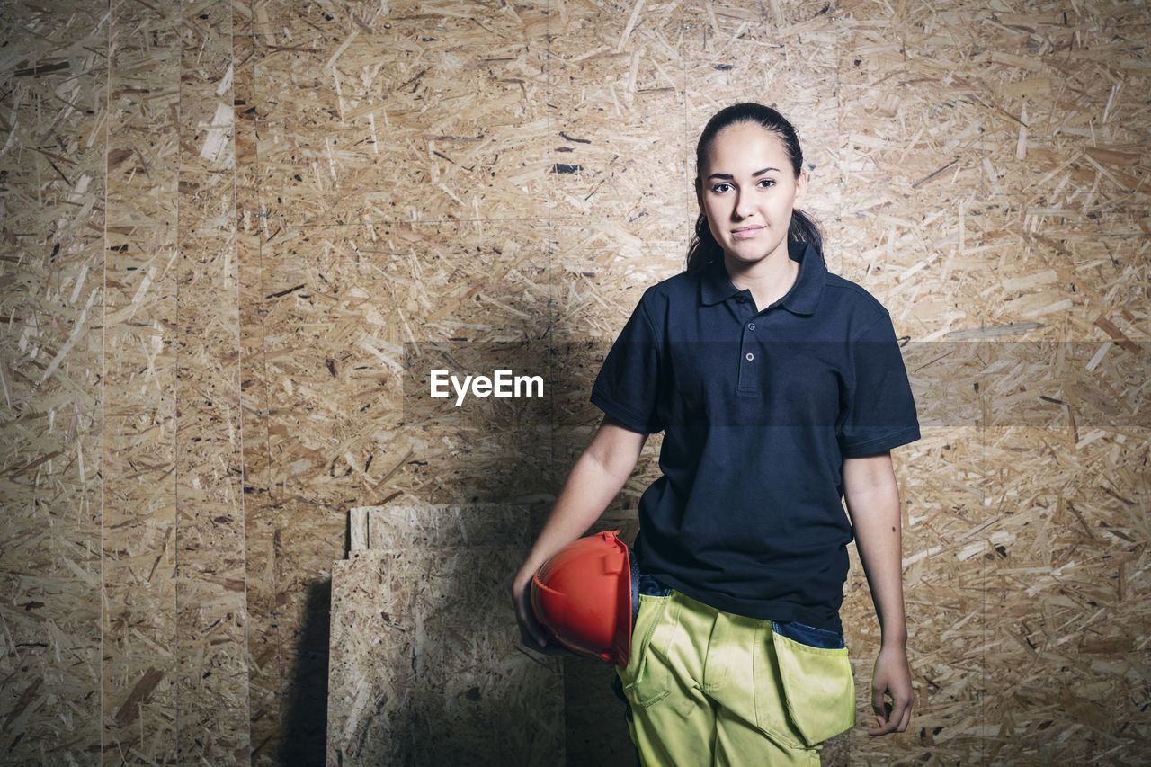 Portrait of confident female carpentry student holding hardhat while standing against wooden wall