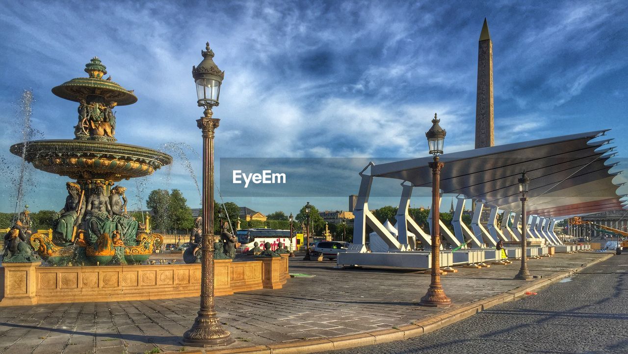 Street lights in front of fountain on sidewalk against cloudy sky