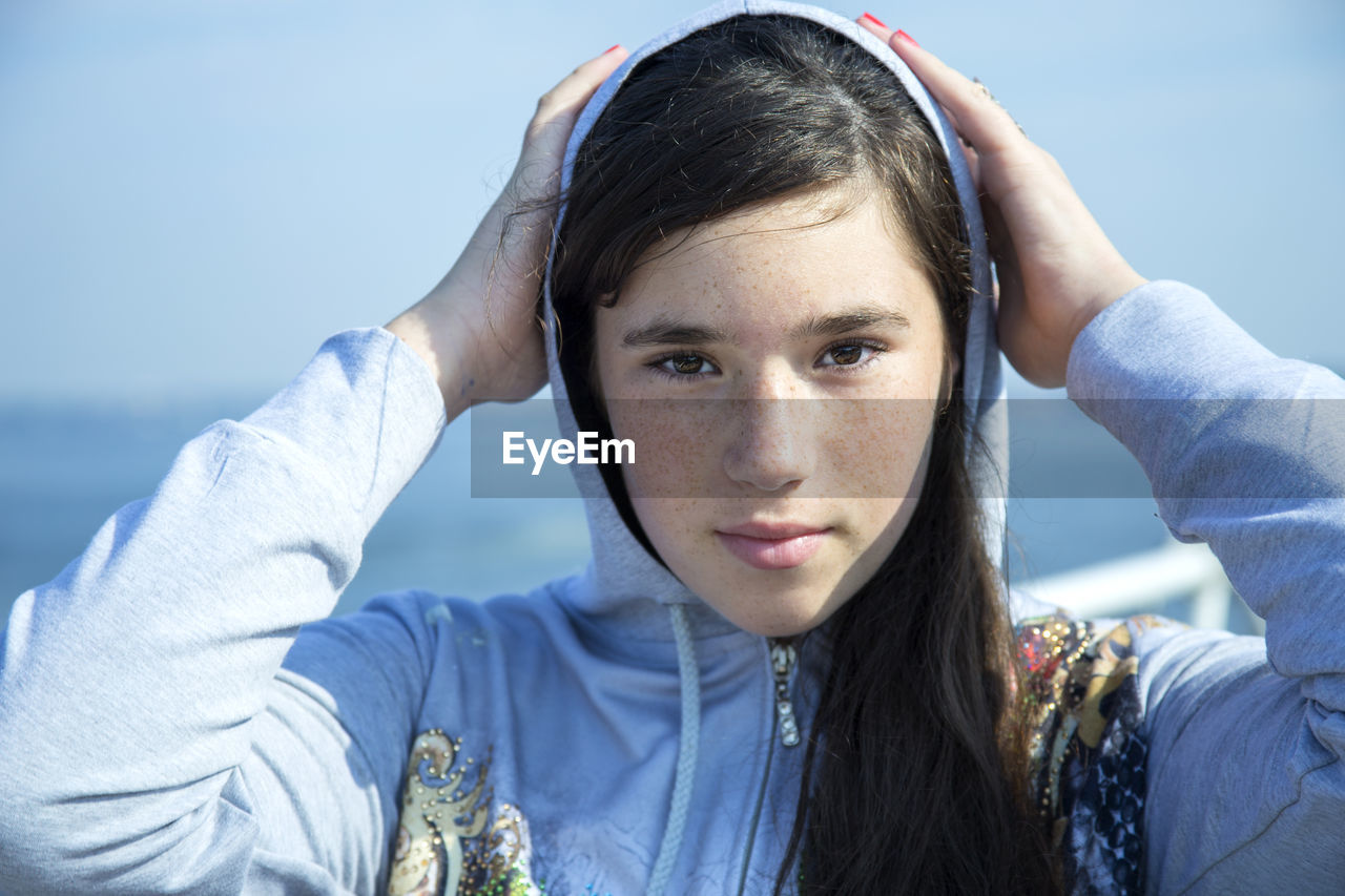 Close-up portrait of young woman in hooded shirt standing against sea