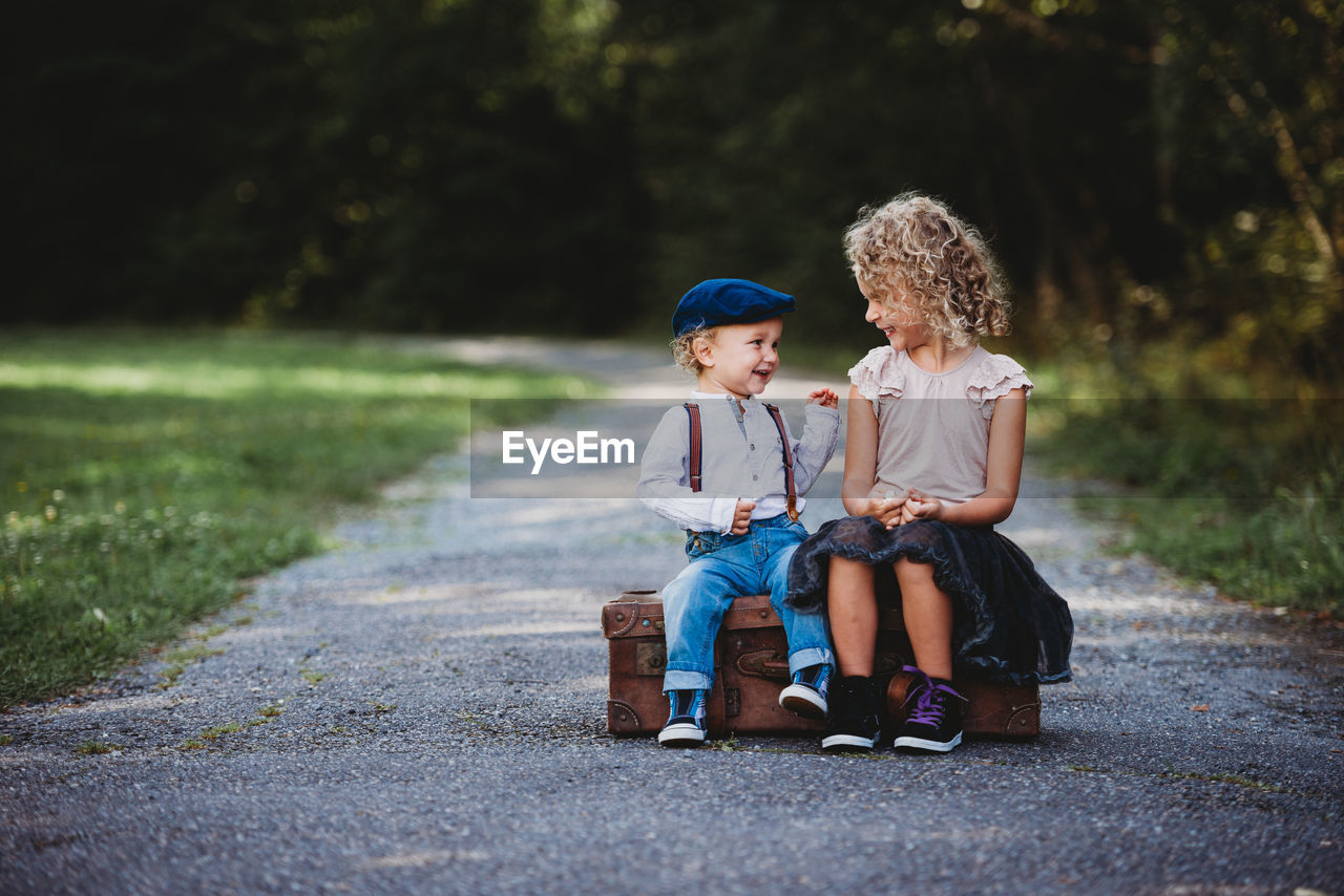 Brother and sister sitting on a vintage suitcase smiling at each other