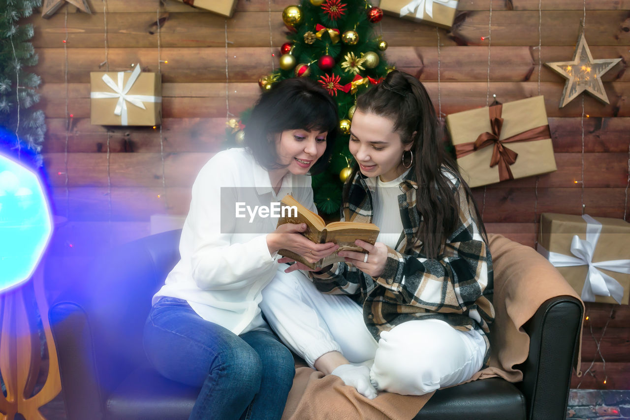 Mom and adult daughter are having fun reading a book on christmas night against the background