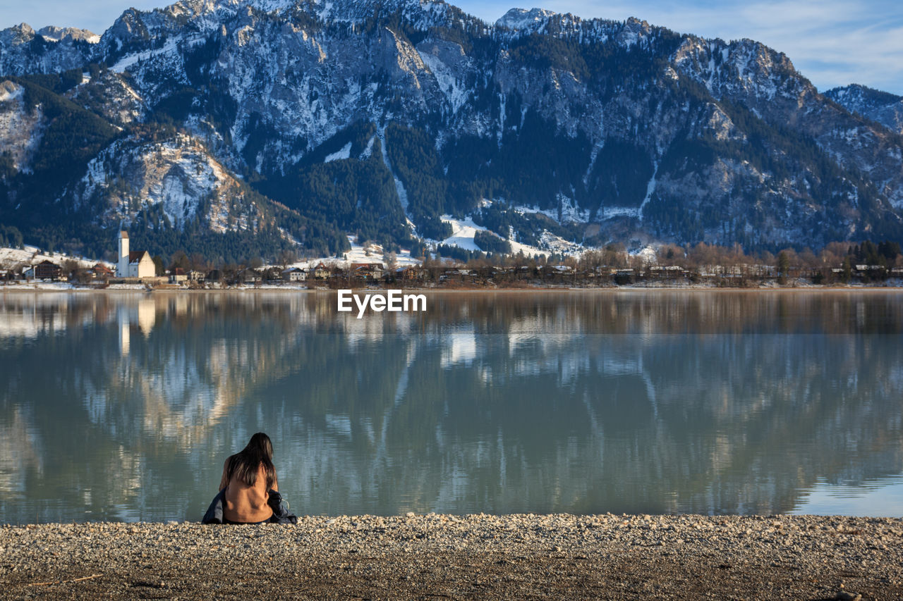 WOMAN SITTING ON ROCK BY LAKE AGAINST MOUNTAIN