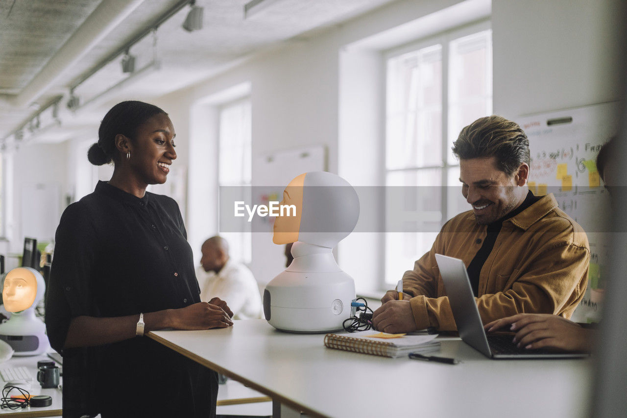 Multiracial smiling students with illuminated social robot at desk in innovation lab