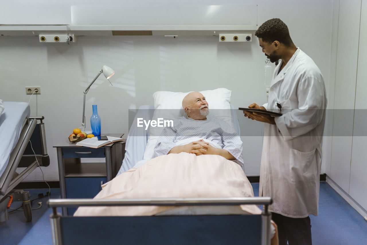 Young male doctor with digital tablet talking to senior patient lying on bed at hospital