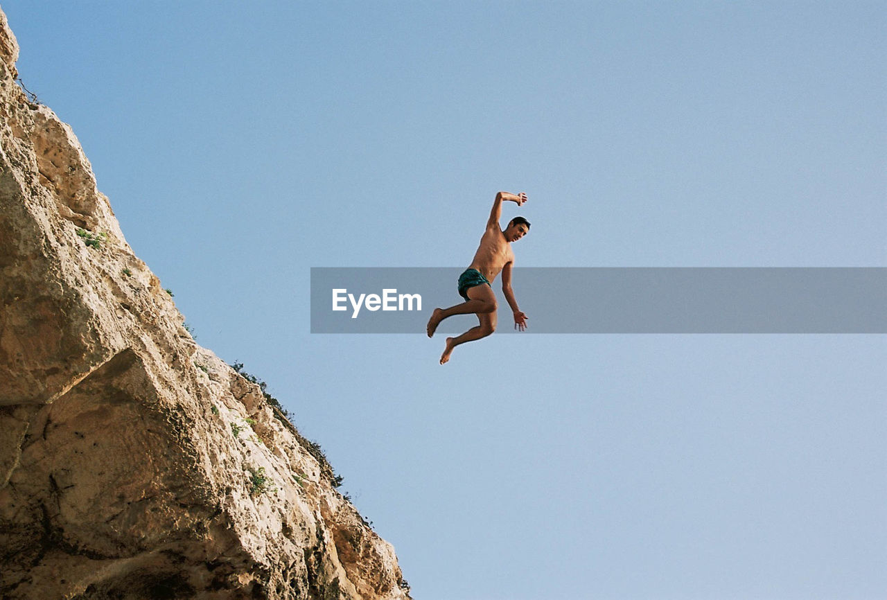 Low angle view of man jumping on rock against clear sky