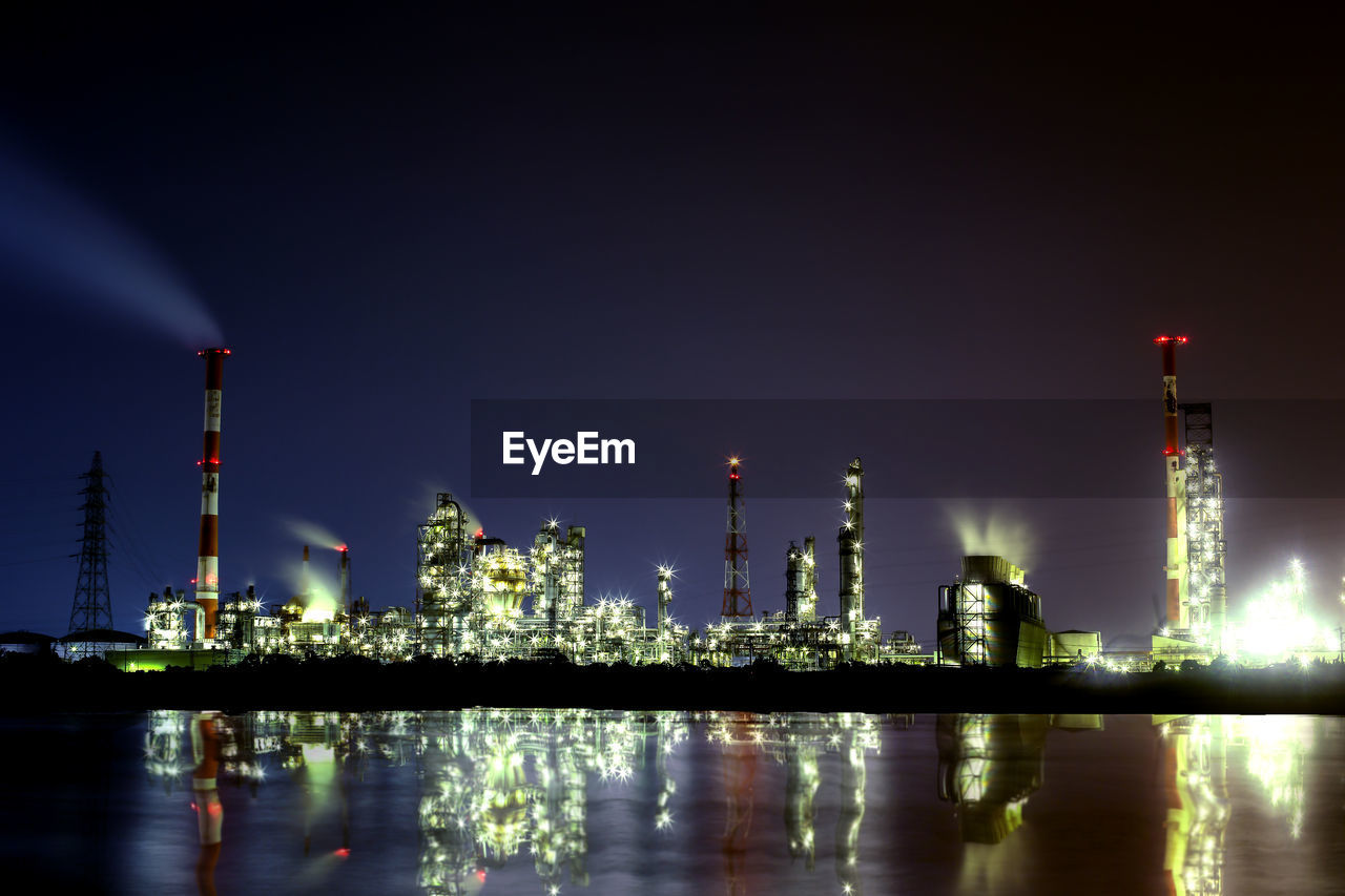 Reflection of illuminated factory on river against sky at night