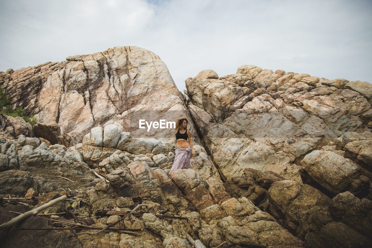 LOW ANGLE VIEW OF WOMAN ON ROCK BY MOUNTAINS AGAINST SKY